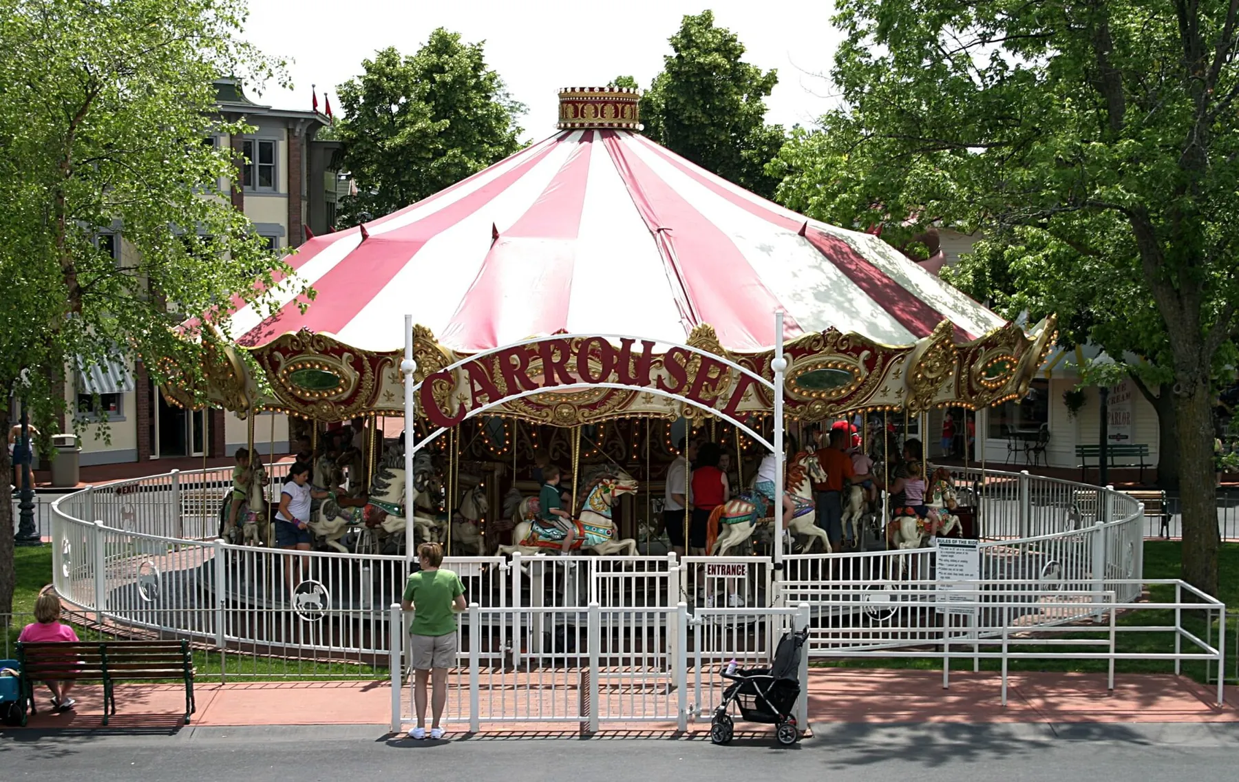 A red-and-white striped carousel with painted horses under shade trees at an amusement park