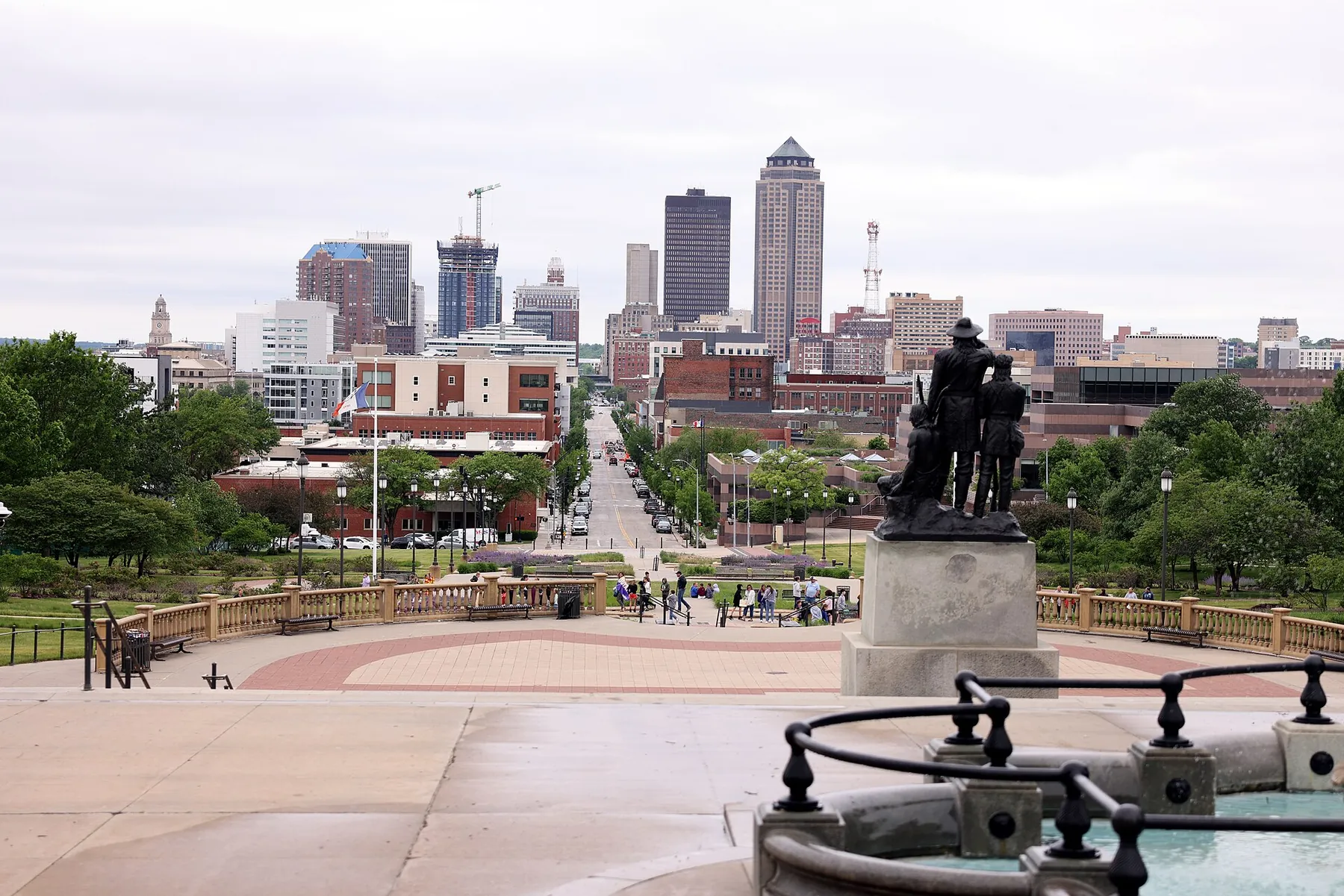 The downtown Des Moines skyline seen from the Iowa State Capitol steps, with a bronze statue and the Capitol plaza in the foreground.